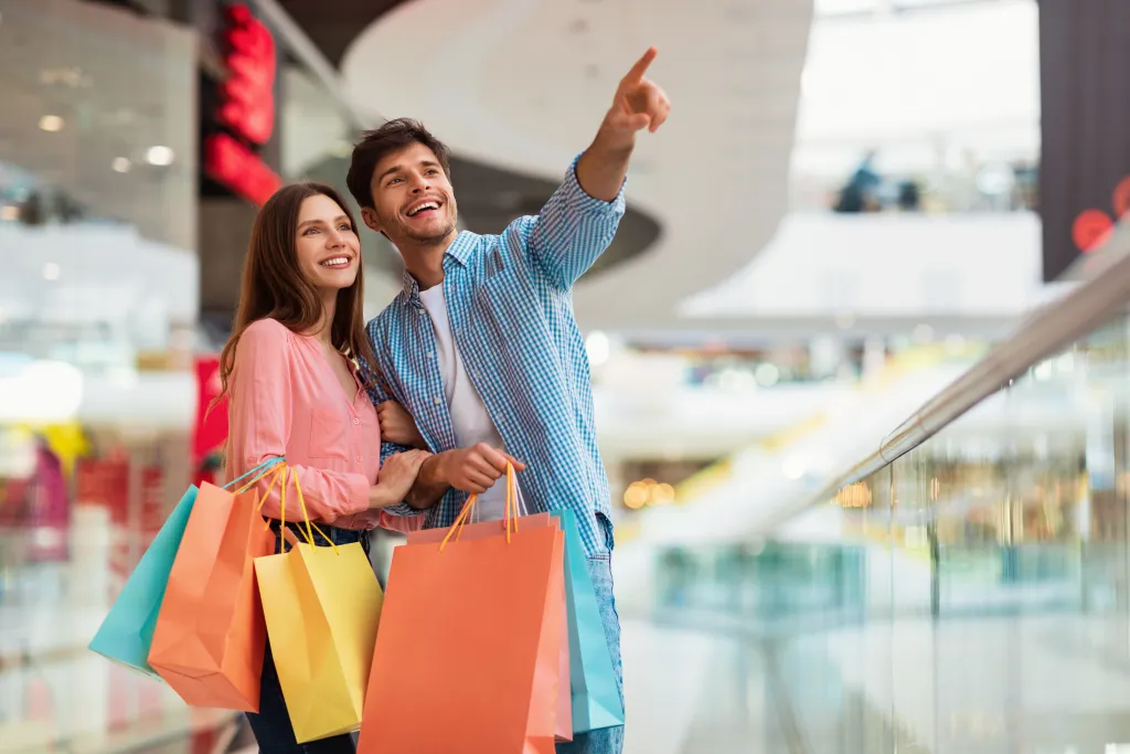 Couple souriant tenant plusieurs sacs de shopping colorés dans un grand centre commercial moderne, l’homme pointant du doigt une boutique ou une attraction au loin.