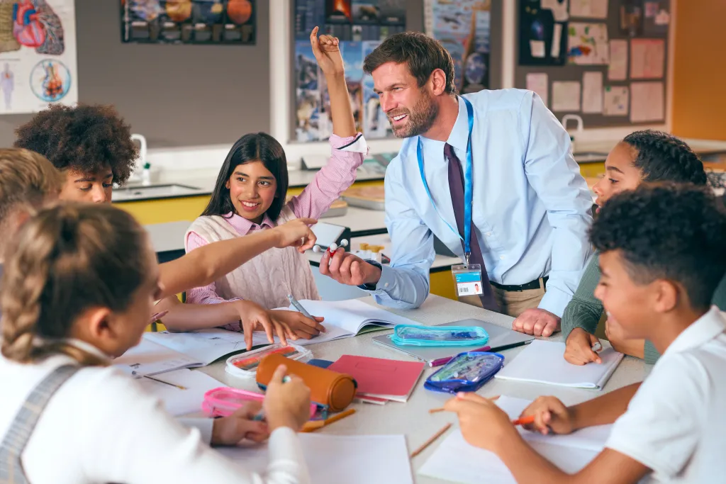 Un enseignant souriant guide un groupe d’élèves aux profils variés pendant qu’ils écrivent et dessinent avec des fournitures colorées dans une salle de classe lumineuse et interactive